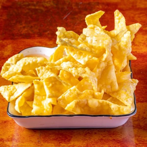 A white bowl filled with crispy, golden-brown wonton strips, likely from Chow n Joy, on a reddish-brown surface.