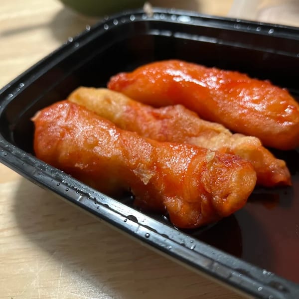 Three crispy, orange-glazed Chinese chicken fingers in a black takeout container with sauce.